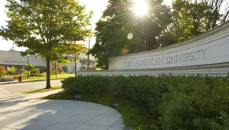 Sun shines through trees near a curved stone sign reading The American University, with green bushes and a building visible in the background.