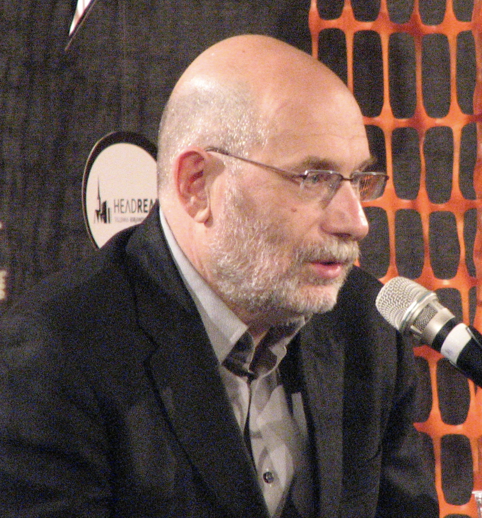 A bald man with glasses and a gray beard, wearing a suit jacket and shirt, speaks into a microphone at an indoor event. An orange safety net and a logoed banner are visible in the background.
