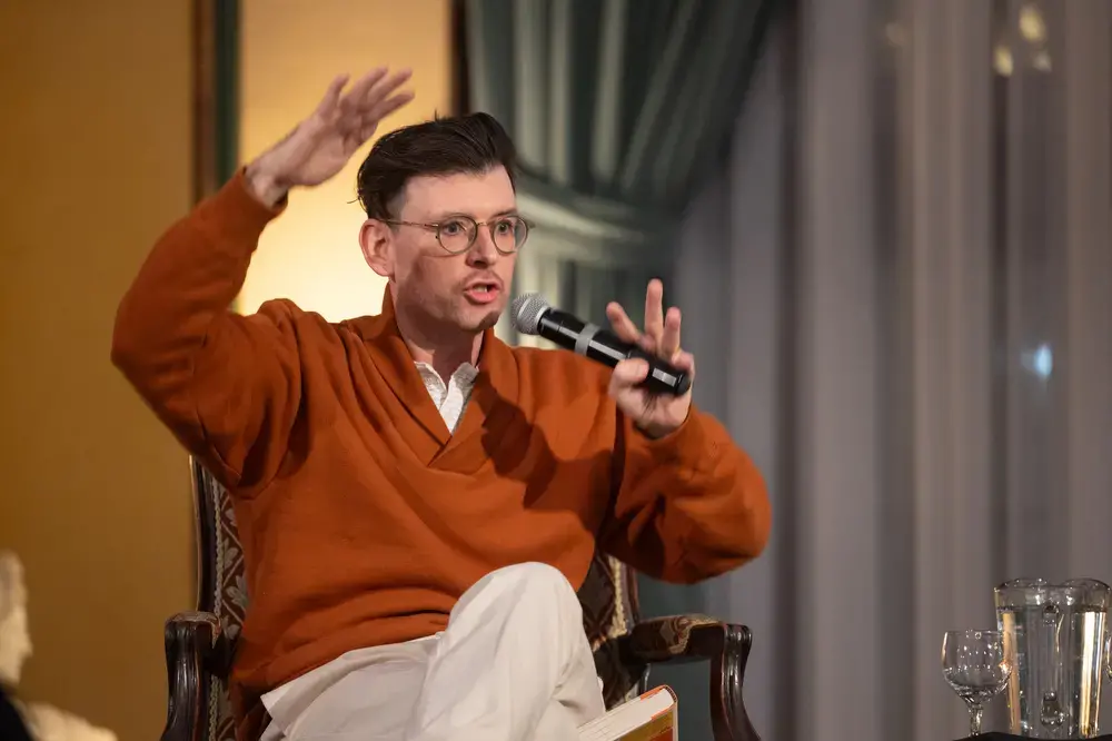 A man in glasses and an orange sweater speaks into a microphone with one hand raised, sitting on a chair during an indoor event. A notebook, water pitcher, and glass are on a table nearby.