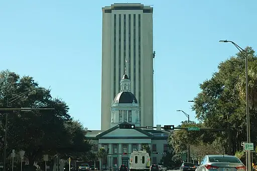A tall government building with a modern tower rising behind a historic structure with a domed roof, surrounded by trees and cars on a sunny day.