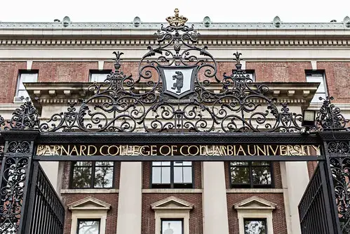 Ornate black iron gate with a crest and gold lettering reading Barnard College of Columbia University, in front of a historic red-brick building with large windows.