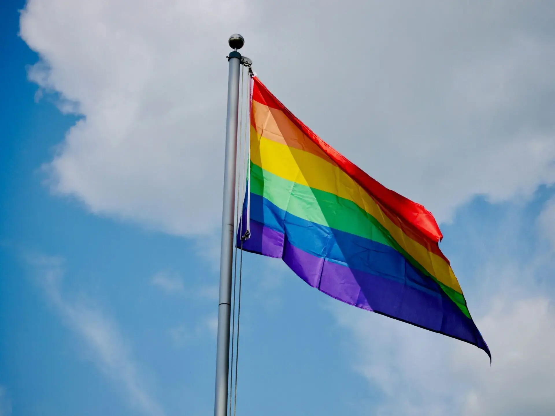 A rainbow pride flag waves on a flagpole against a partly cloudy sky, symbolizing LGBTQ+ pride and diversity.