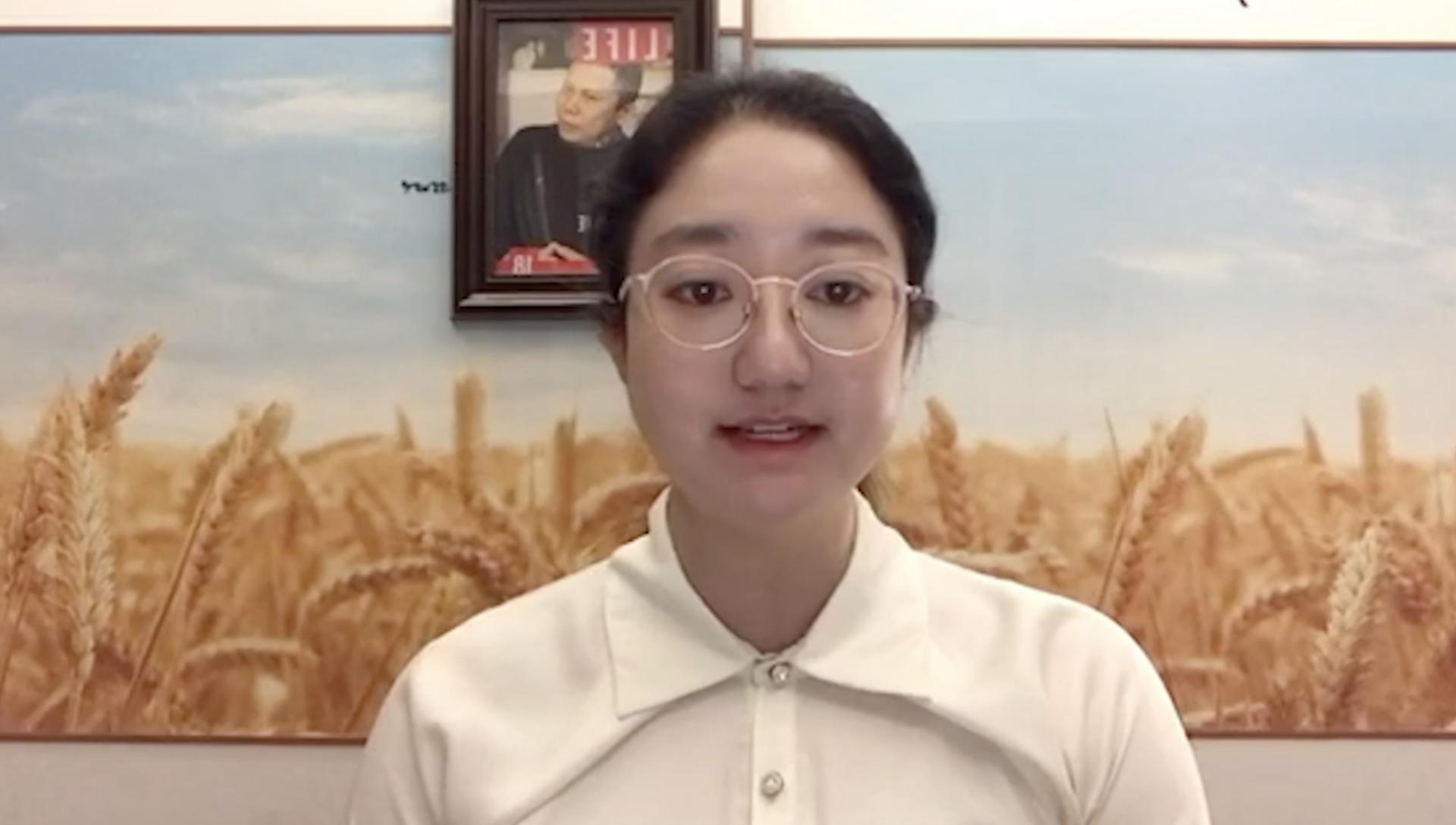 A woman with glasses and a white collared shirt sits in front of a backdrop featuring wheat fields. A framed picture of a person hangs on the wall behind her.