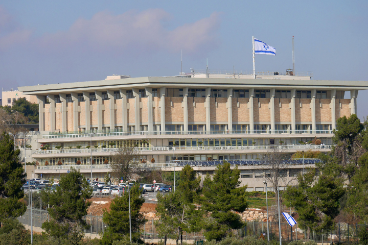The Knesset building in Jerusalem, Israel, with an Israeli flag flying on top and surrounded by trees and parked cars on a clear day.
