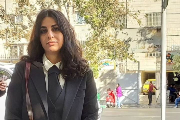 A woman with long dark hair, wearing a black suit, white shirt, and black tie stands outdoors. Behind her are trees, parked vehicles, and a sign with Arabic writing. It is a sunny day.