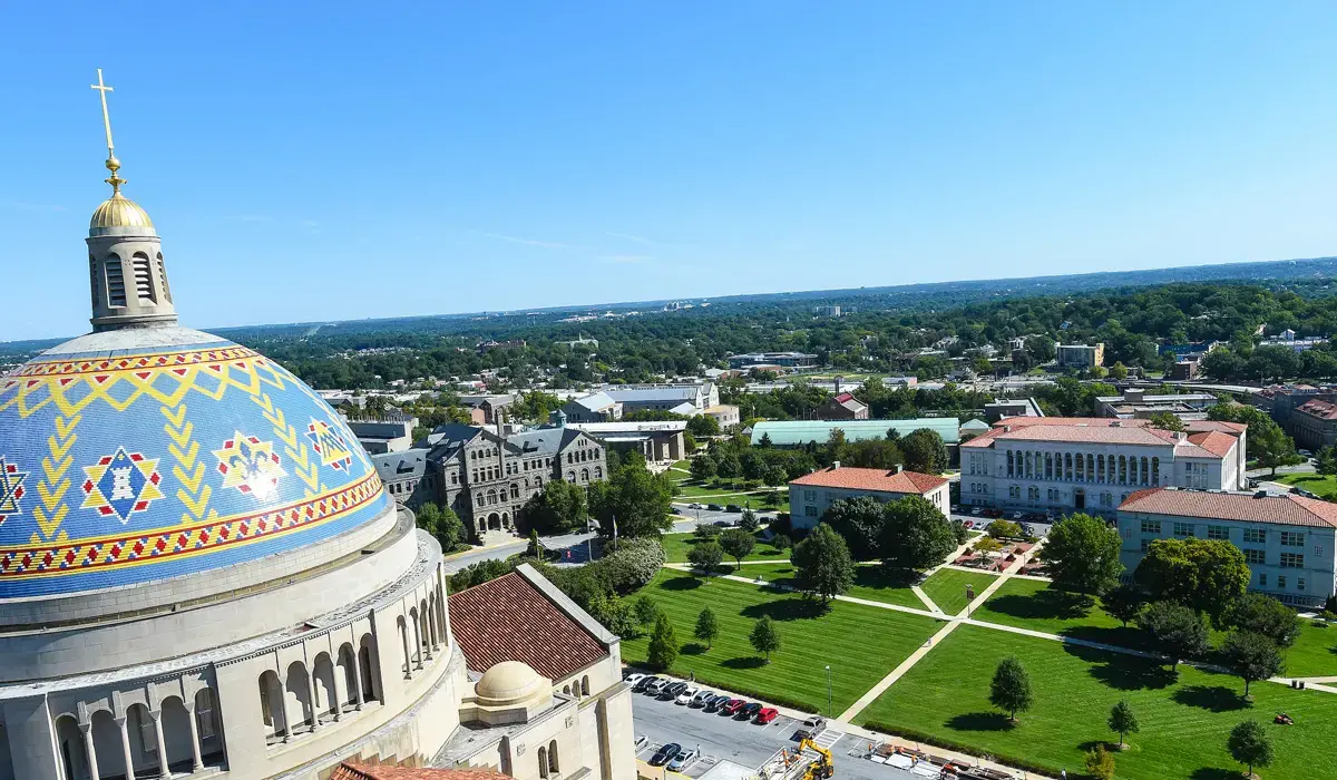Aerial view of a university campus with green lawns, red-roofed buildings, and a large domed church featuring colorful mosaic patterns under a clear blue sky.