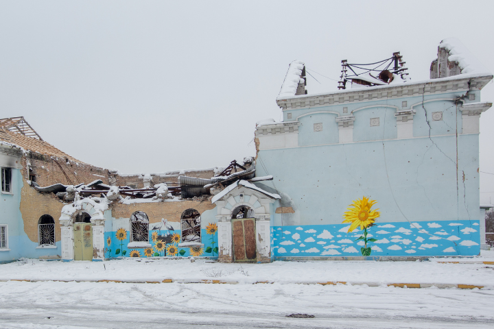 A partially destroyed building in winter, with snow covering the ground and roof. The remaining wall is painted with yellow sunflowers and white clouds on a blue background, creating a stark contrast with the damage.