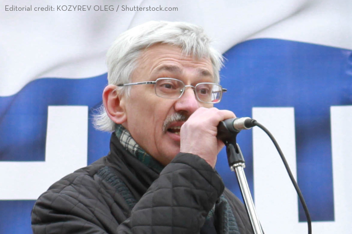 An older man with gray hair, glasses, and a mustache speaks passionately into a microphone at an outdoor event. He wears a scarf and quilted jacket. A blurred blue and white banner is visible in the background.