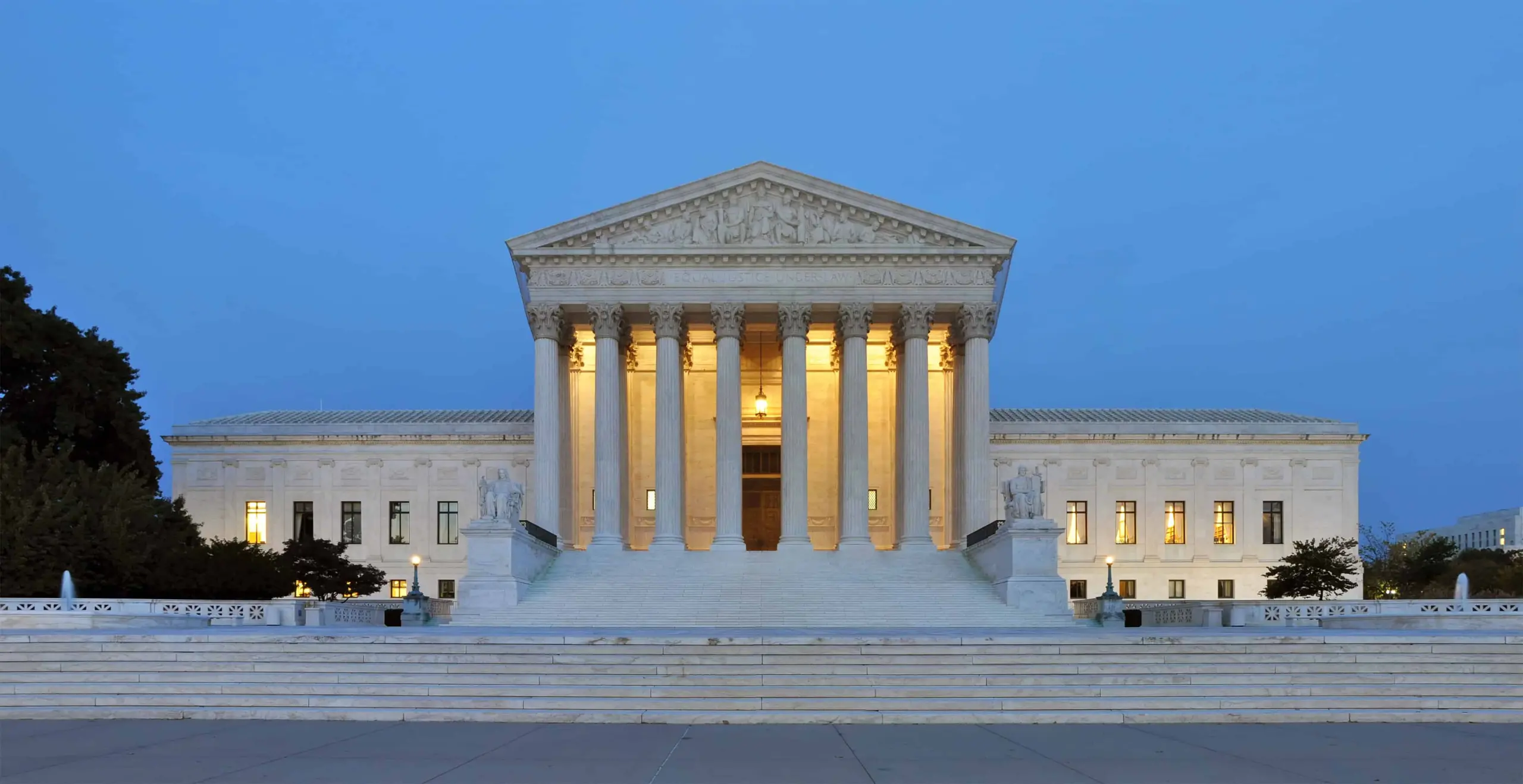 The United States Supreme Court building at dusk, with its grand columns and steps illuminated by warm lights against a clear blue sky.