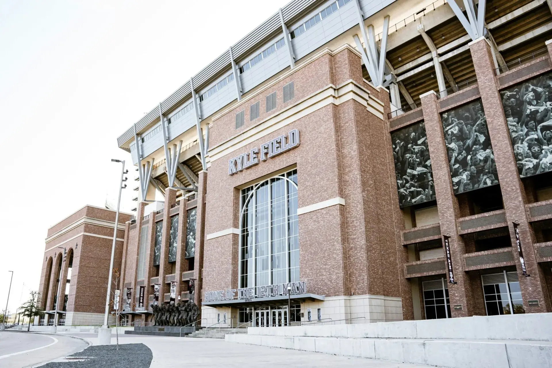 A wide view of Kyle Field stadium’s exterior, showing red brick walls, tall glass entrance doors, large windows, and metallic structural beams, with statues in front and the stadium name displayed above the entrance.