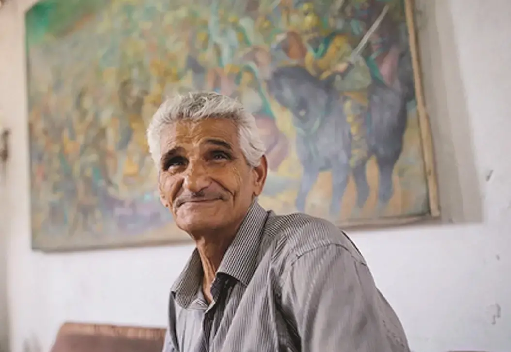 An older man with gray hair and a striped shirt smiles gently, sitting indoors. A colorful, blurry painting of a battle scene hangs on the wall behind him.