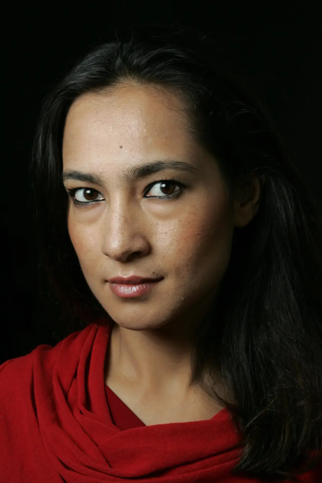 A woman with long dark hair and brown eyes looks into the camera, wearing a red top against a dark background. Her expression is neutral and confident.