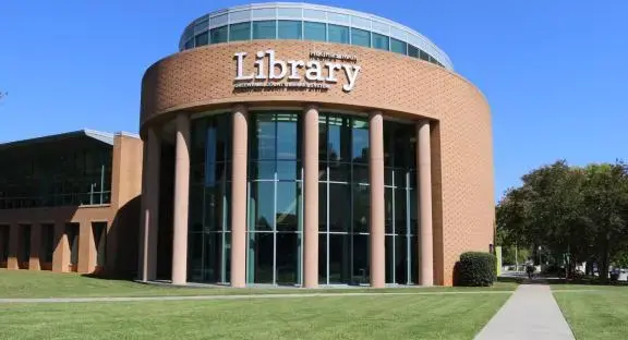 Exterior of a modern library with a circular brick facade and tall glass windows, featuring large columns. A curved pathway leads through a grassy lawn surrounded by trees, under a clear blue sky.