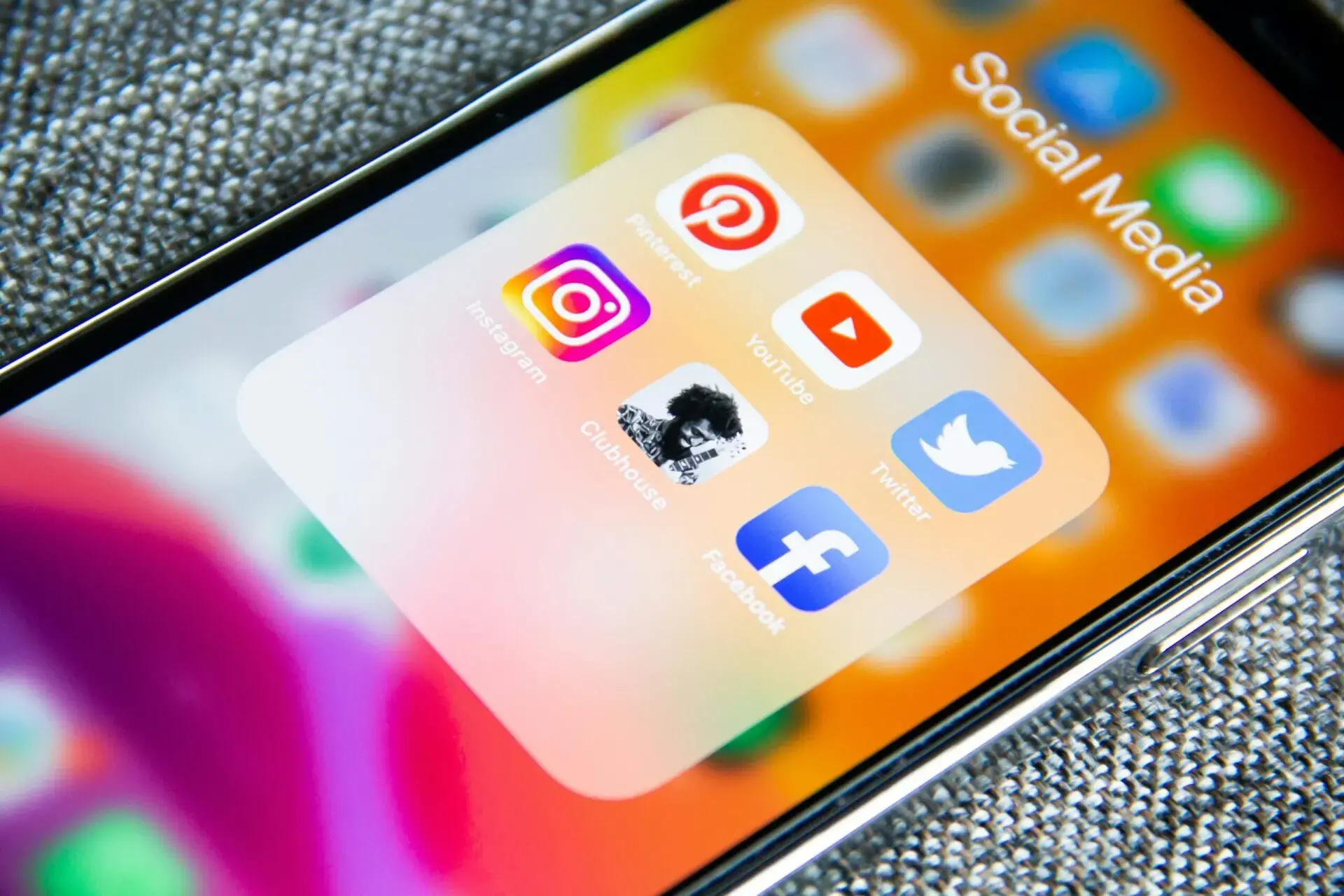 A smartphone screen displays a folder labeled Social Media containing the Pinterest, YouTube, Instagram, Clubhouse, Twitter, and Facebook app icons. The background is colorful and slightly blurred.