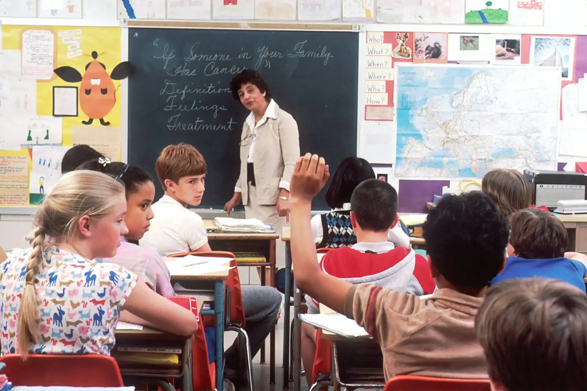 A classroom scene shows students seated at desks facing a teacher at the chalkboard. One student raises their hand, while others look toward the teacher. The board has writing, and a map of Europe is visible on the wall.