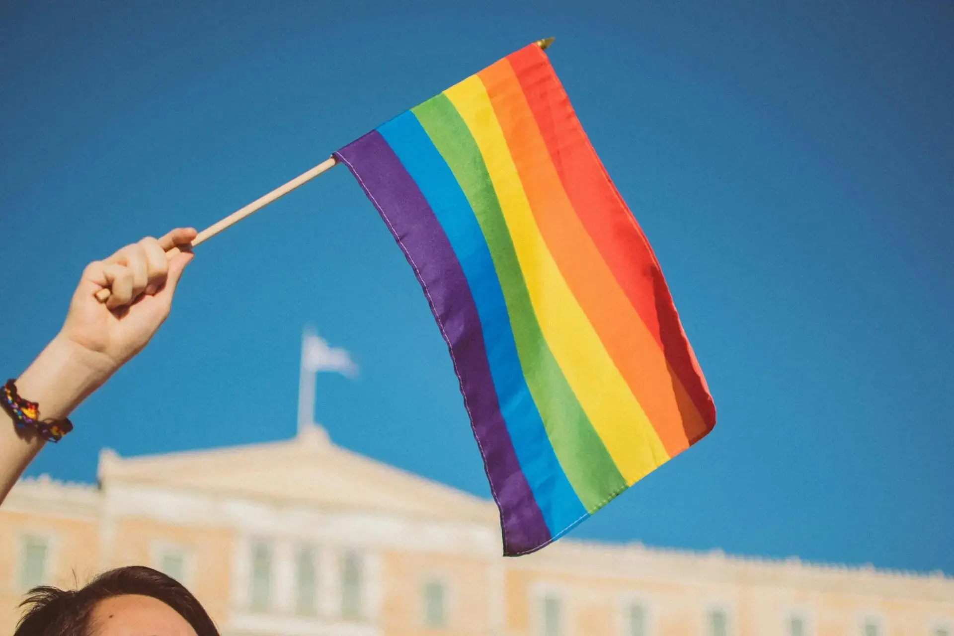 A hand holds up a rainbow pride flag against a clear blue sky, with a building and a flag in the background.