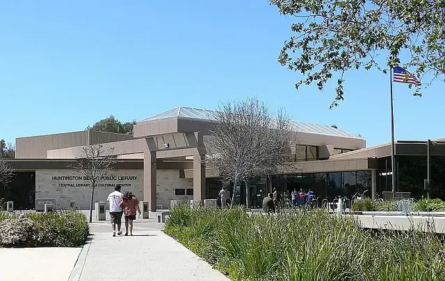 People walk toward a modern, beige building labeled “Huntington Beach Public Library” on a sunny day. There are trees, greenery, and an American flag on a pole outside the entrance.