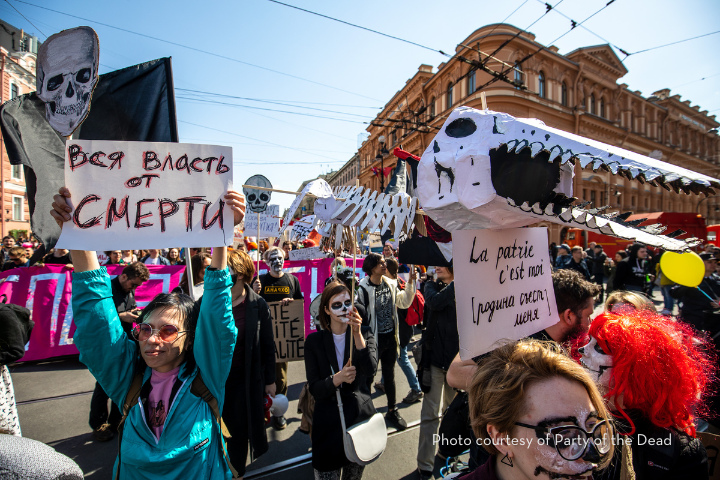 A crowd marches down a city street, many wearing skull face paint and holding large skull puppets and protest signs in Russian and French. Historic buildings line the street under a clear, sunny sky.