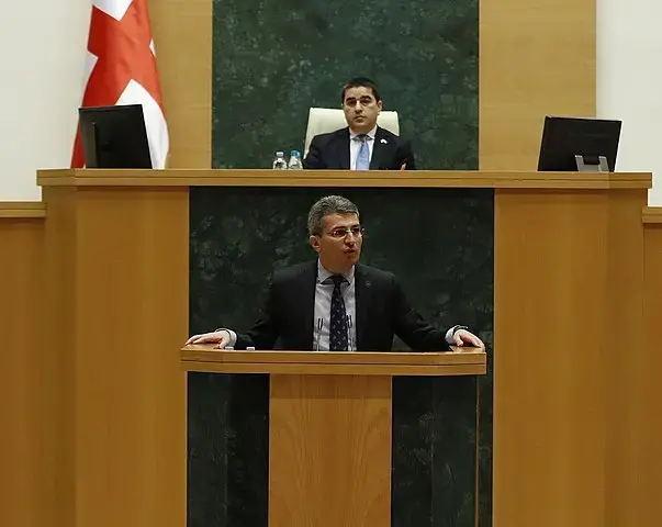 A man in a suit speaks at a podium in a parliamentary chamber, while another man sits behind him at a raised desk. A large flag with red and white colors is partially visible on the left.