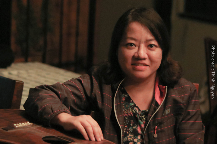 A woman with shoulder-length dark hair, wearing a striped jacket over a floral top, sits indoors and smiles at the camera with her hand resting on a wooden surface. Photo credit: Thinh Nguyen. The scene captures Pham Doan Trang in a moment of warmth and ease.