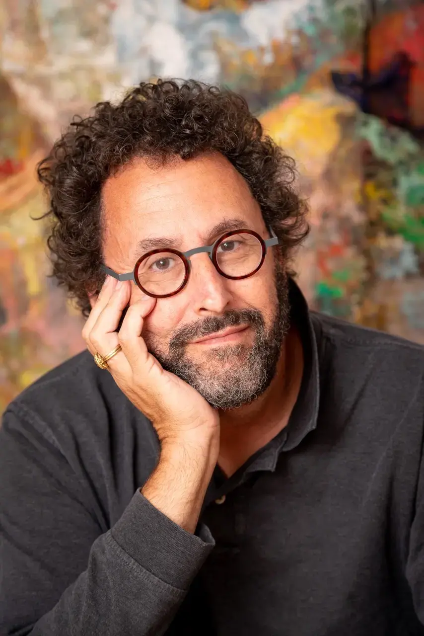 A man with curly dark hair, a beard, and glasses rests his face on his hand, wearing a dark shirt, sitting in front of a colorful, abstract background.