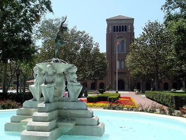A stone fountain with statues and water sits in a courtyard, with a brick building, gardens, and an American flag in the background on a sunny day.