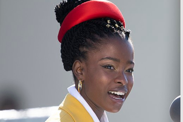 A young Black woman with braided hair, resembling Amanda Gorman, wears a bright red headband, gold hoop earrings, and a yellow blazer over a white shirt as she speaks or sings into a microphone outdoors.
