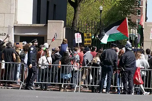 A crowd gathers behind metal barricades at a protest. People hold signs and flags, including a prominent Palestinian flag. Police officers stand nearby, monitoring the scene. Trees and a building are visible in the background.