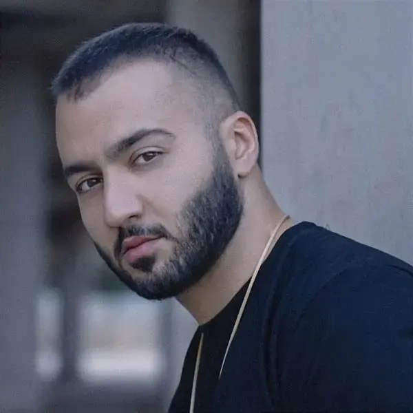 A man with short hair and a full beard, wearing a black shirt and a gold chain, looks seriously at the camera against a blurred neutral background.