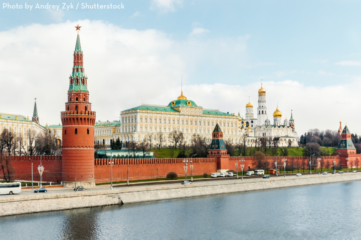 View of the Moscow Kremlin with its red brick walls, towers, and golden-domed cathedrals beside the Moskva River, under a partly cloudy sky.