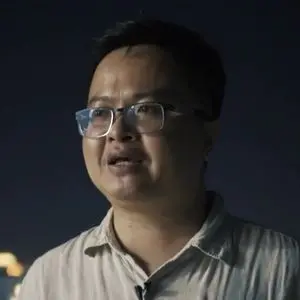 A person with short dark hair and glasses is standing outdoors at night, wearing a light-colored collared shirt. The background is dark with blurred lights visible.
