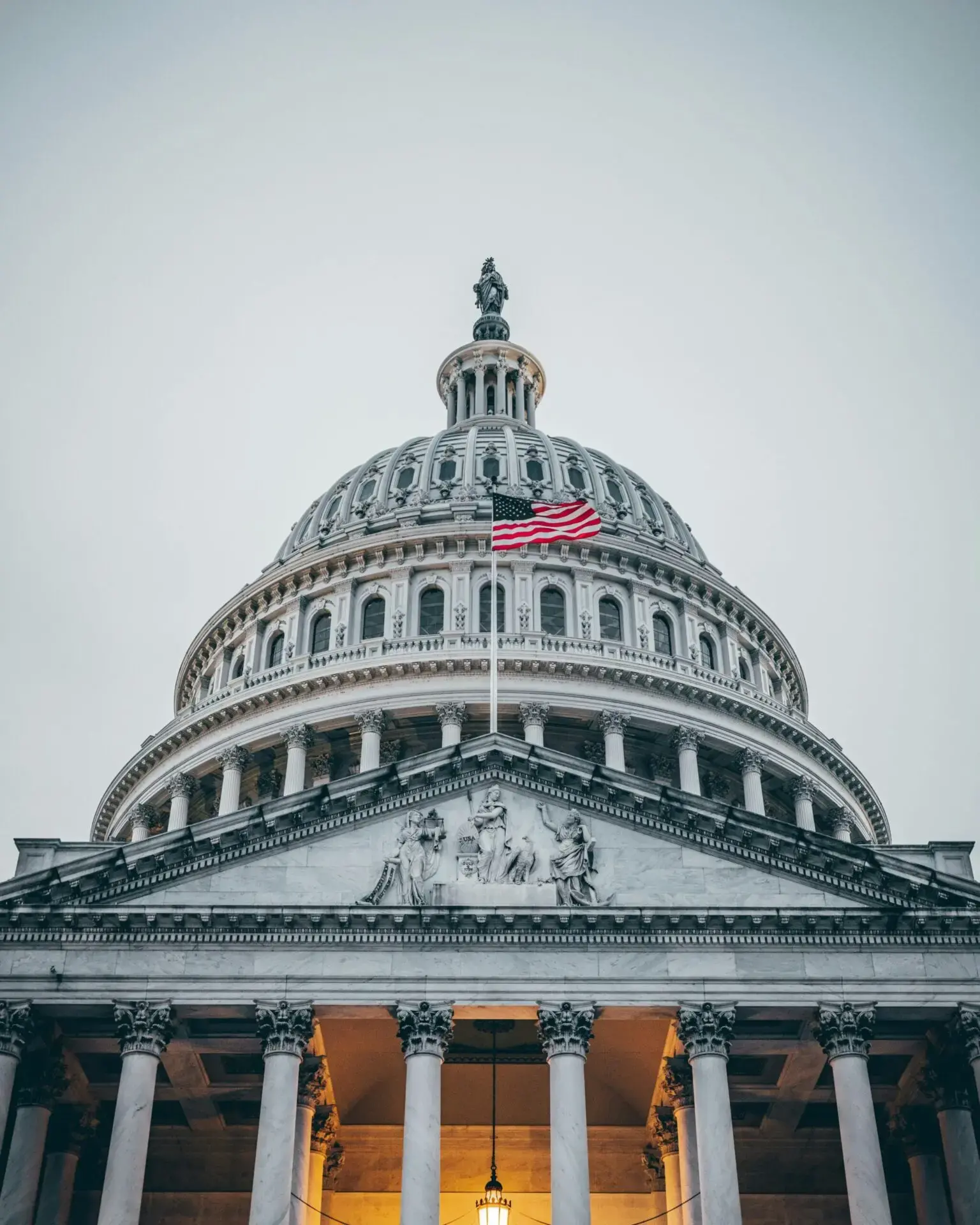 The United States Capitol building’s dome is seen from below, with an American flag flying in front. The sky is overcast, and the building’s columns and detailed carvings are visible.