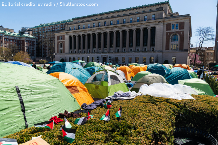 Dozens of colorful tents are set up on a university lawn in front of a large academic building, with small Palestinian flags visible in the foreground. The scene depicts a protest encampment.