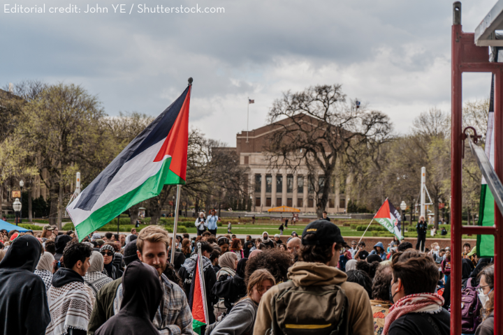A crowd gathers outdoors holding Palestinian flags, with a large academic building and leafless trees in the background. The sky is overcast, and people are dressed in jackets and scarves.