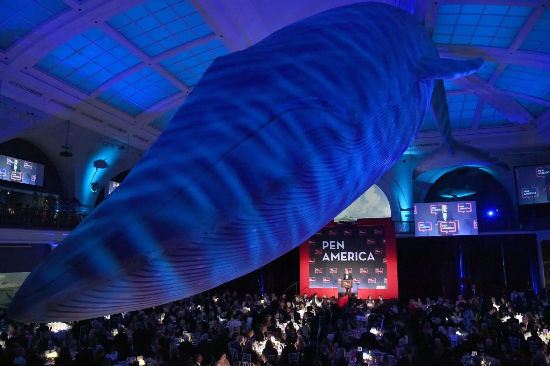 A large blue whale model hangs from the ceiling over a crowded event with banquet tables and a red PEN America news stage backdrop, all under dramatic blue lighting.