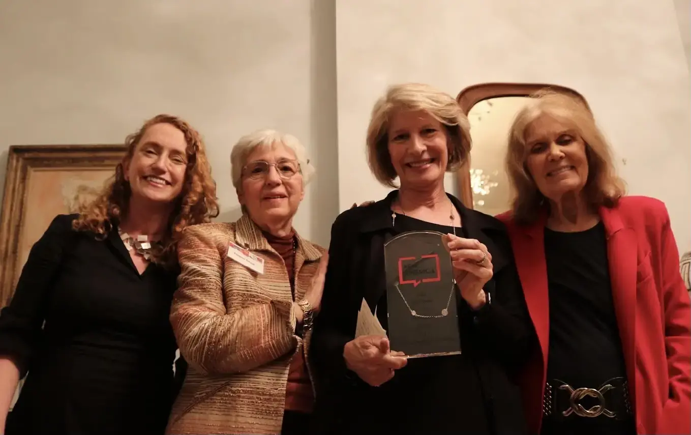 Four women stand together smiling indoors. The woman second from the right holds an award shaped like a black plaque with a red outline of a computer screen. They appear to be attending a formal or celebratory event.