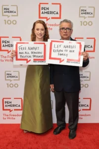 A woman and a man stand on a red carpet holding signs that read “Free Narges and all Iranian political prisoners” and “Let Narges return to her family” at a PEN America event, highlighting that Narges Mohammadi was awarded the Nobel Peace Prize.