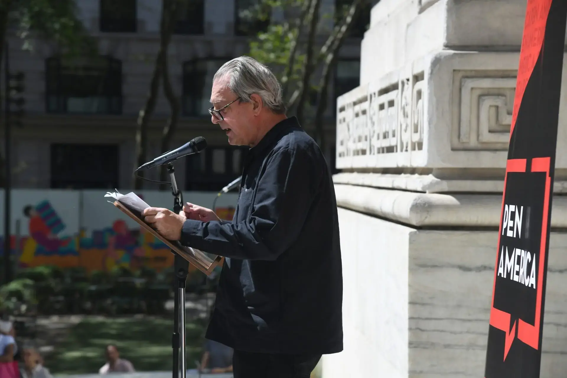 A person wearing glasses and a dark shirt reads from papers into a microphone at an outdoor event, standing beside a stone building. A PEN AMERICA sign is visible on the right; the scene evokes readings by authors like Paul Auster.
