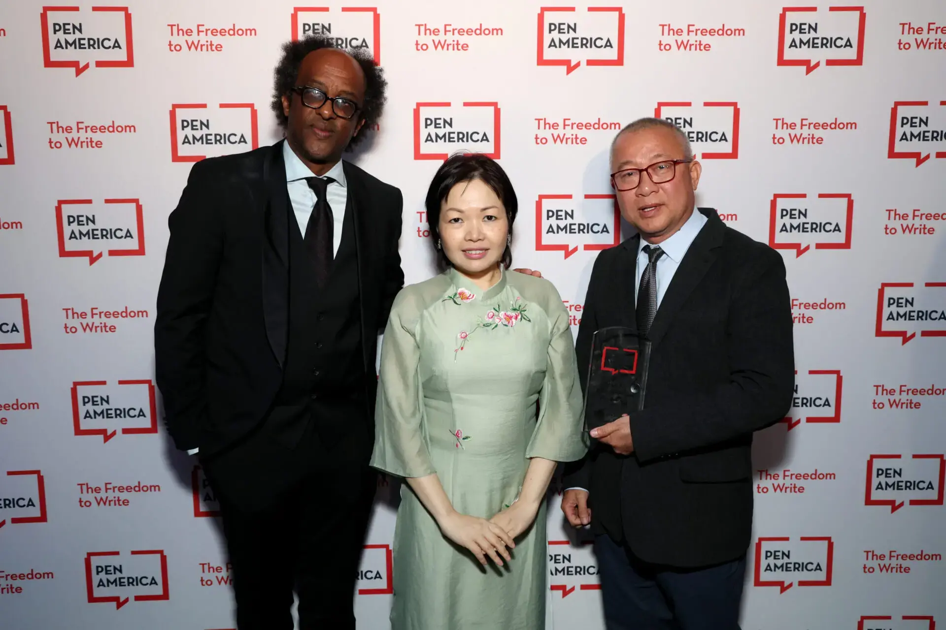 Three people stand together in front of a backdrop with PEN America and “The Freedom to Write” logos. The two men wear suits and the woman wears a light green dress with floral embroidery. One man holds an award plaque.