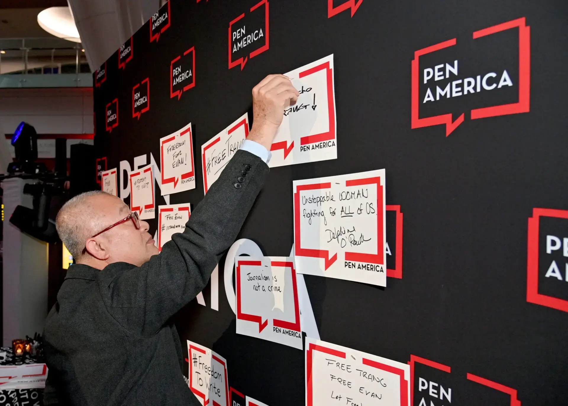 A person writes a message on a note and attaches it to a wall filled with similar notes at a PEN America event. The wall and the notes prominently display the PEN America logo.