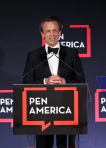 A man in a tuxedo stands at a podium with a PEN AMERICA sign, speaking into microphones at an event. The background features the PEN America logo repeatedly.