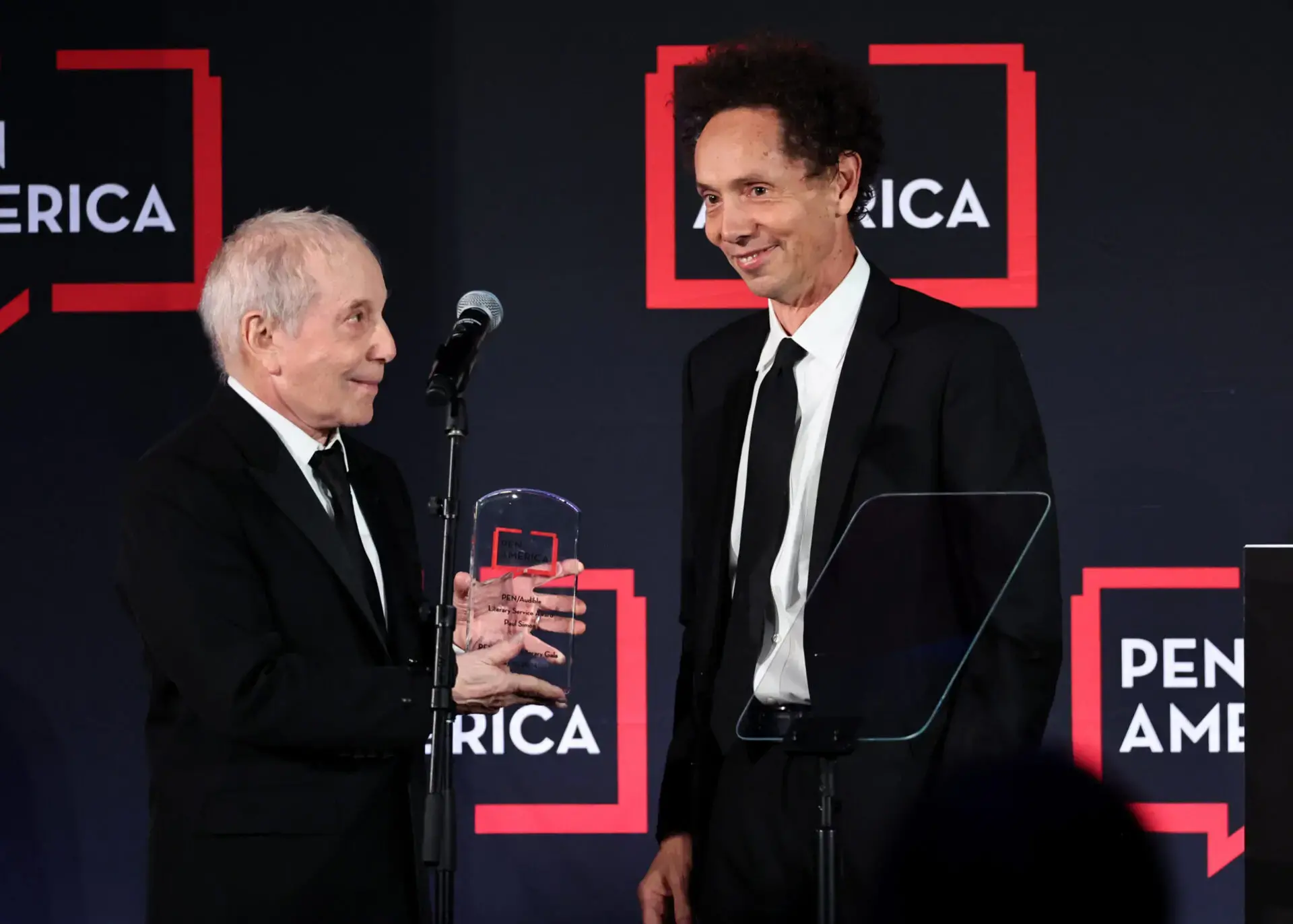 Two men in suits stand on stage at a PEN America event. One hands the Paul Simon Award to the other while they both smile. A microphone and podium are in front of them, with the PEN America logo visible in the background.