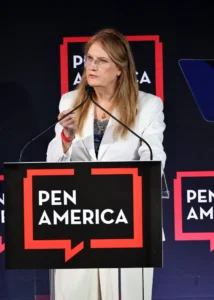 A woman in a white suit speaks at a podium with a PEN America sign, gesturing with her hand. The backdrop features the PEN America logo.