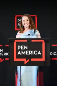 Suzanne Nossel stands at a podium with a PEN America sign, smiling in her light blue dress against a black background decorated with red and white PEN America logos at the gala.