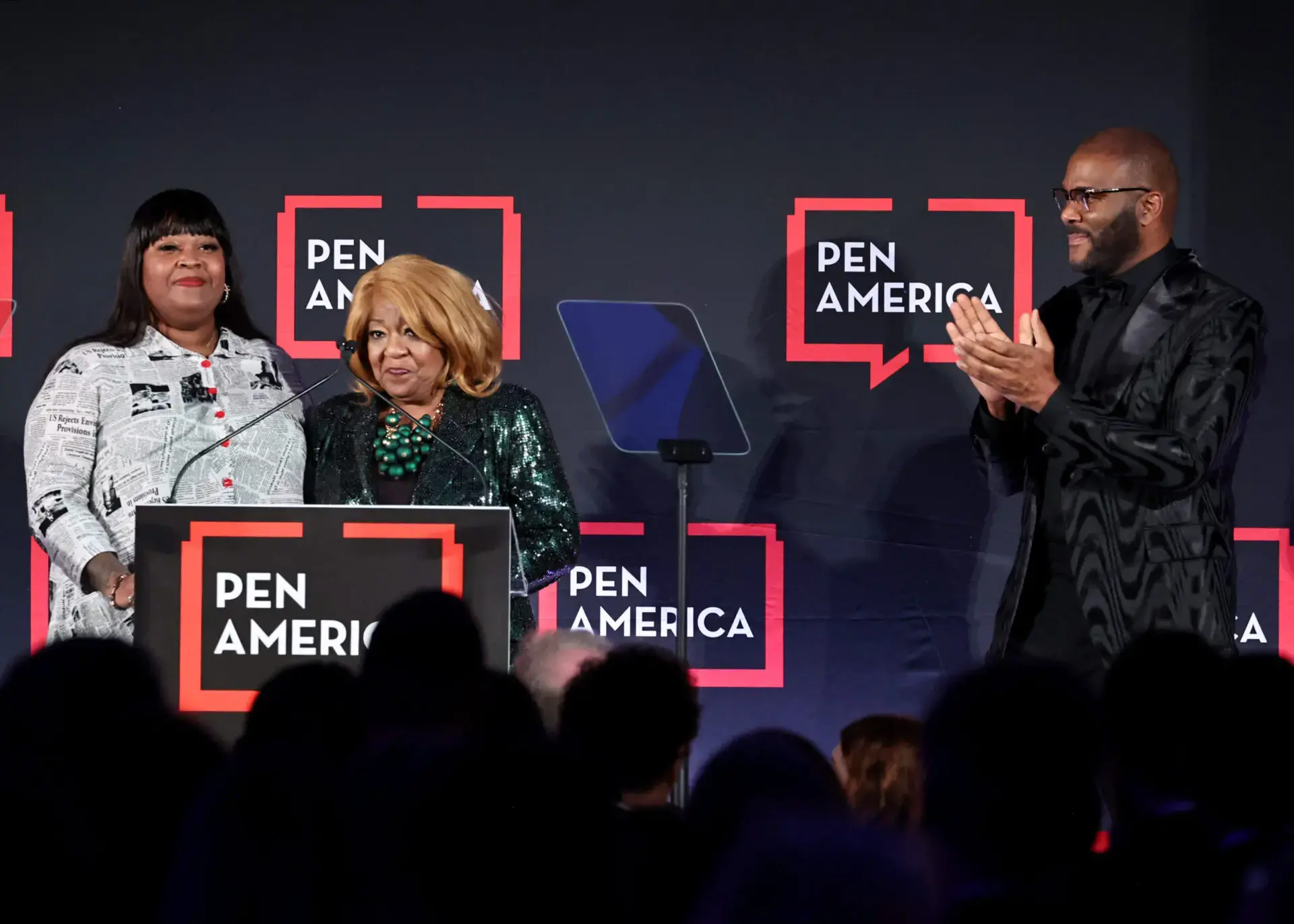 Three people stand on stage at a PEN America event; two women are at a podium, one speaking, while a man on the right side of the image stands and applauds. The PEN America logo appears repeatedly in the background.