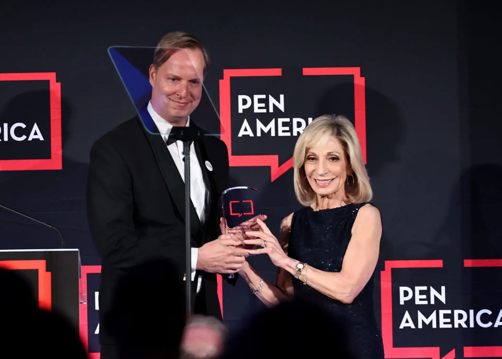 A man in a tuxedo and a woman in a navy dress smile as they hold an award together on stage at a PEN America event, with the PEN America logo in the background.