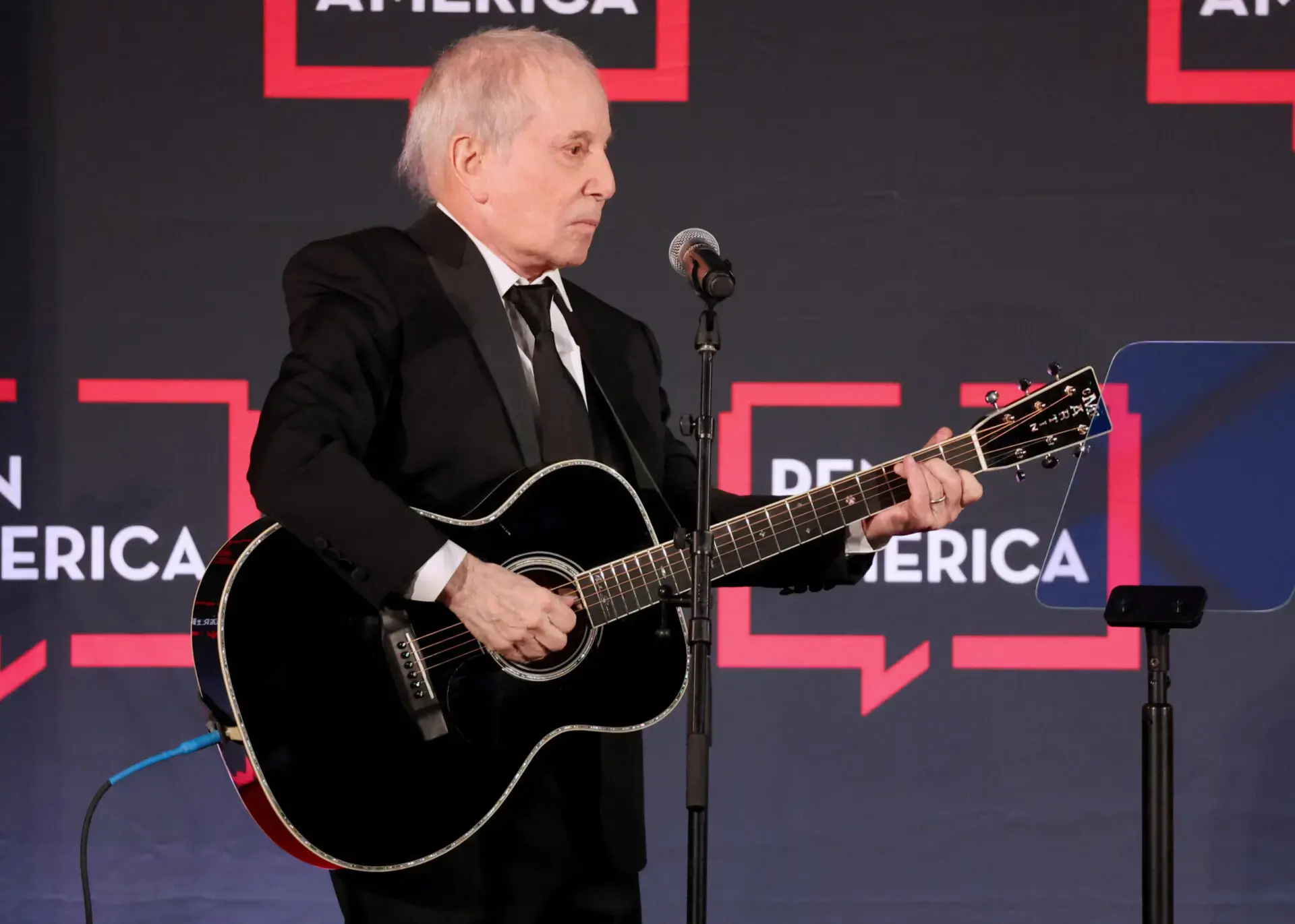 An older man in a black suit plays a black acoustic guitar and sings into a microphone on stage, with a backdrop featuring red and white text and speech bubble graphics.