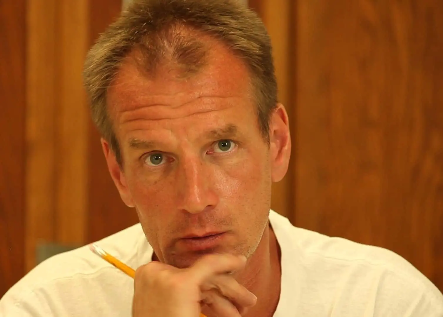 A man with short brown hair and blue eyes, wearing a white shirt, holds a pencil near his chin and looks thoughtfully at the camera. The background is wooden paneling.
