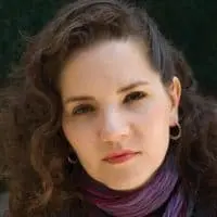 A woman with long curly brown hair, wearing hoop earrings and a purple scarf, looks directly at the camera with a neutral expression. The background is dark and blurred.