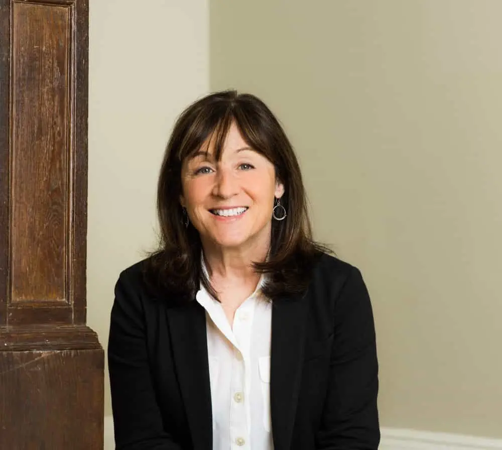 A woman with shoulder-length brown hair, wearing a black blazer over a white shirt, sits indoors and smiles at the camera. There is a wooden pillar beside her and a plain light-colored wall in the background.